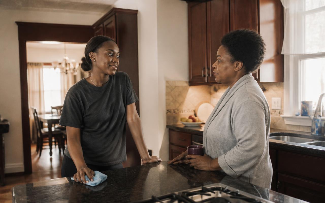 A housekeeper and a parent talking across a kitchen counter in warm morning light